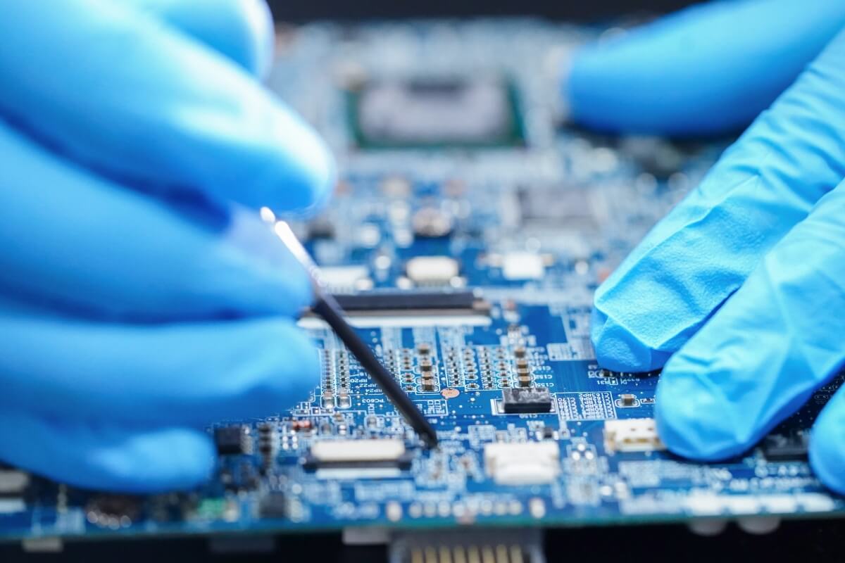 Two gloved hands on top of a printed circuit board. One hand is holding a tool, preparing to perform conformal coating removal.