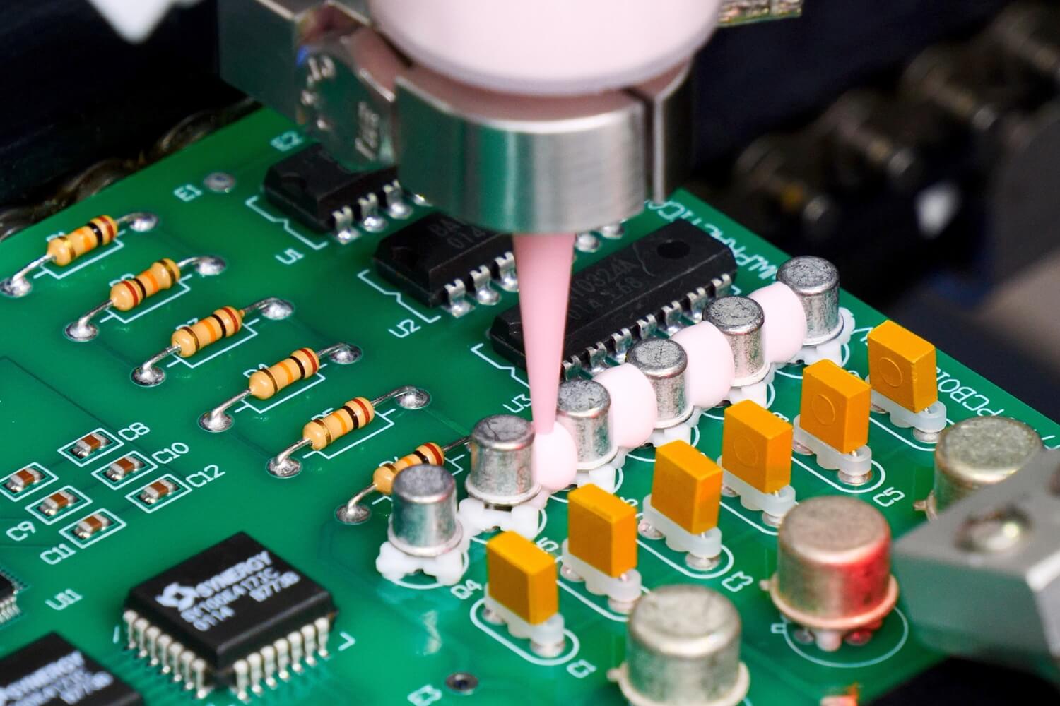 Close-up of the SCS PrecisionCoat VI using a SCS dispense valve to apply thick light pink coating material around silver components on a green circuit board.