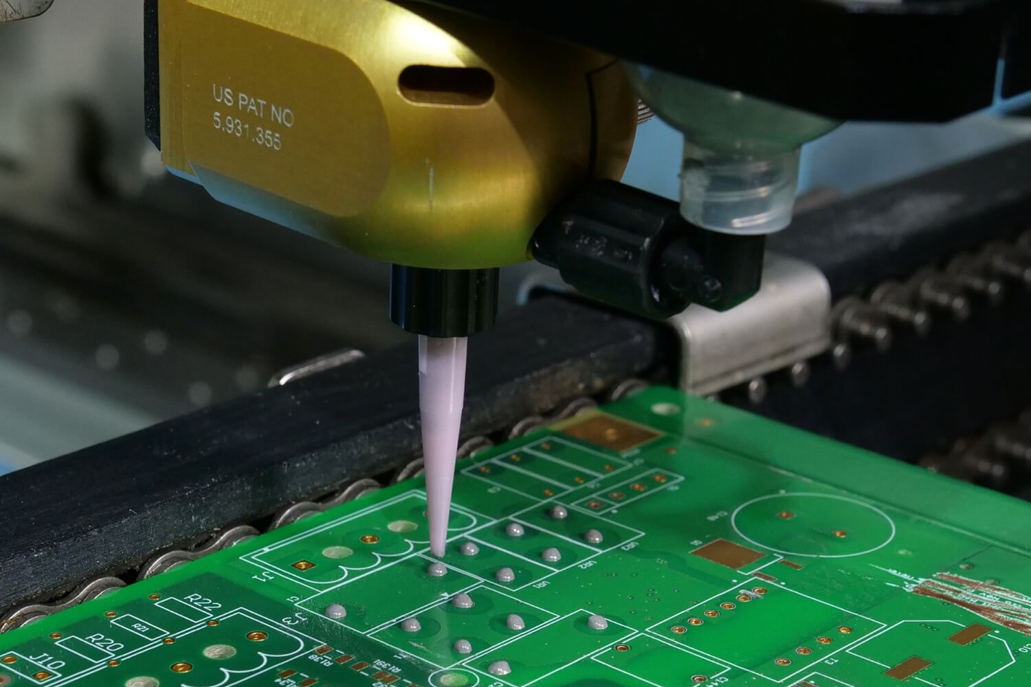 Close-up of the SCS PrecisionCoat VI using an auger valve to apply gray coating material in a dot formation onto a green circuit board.