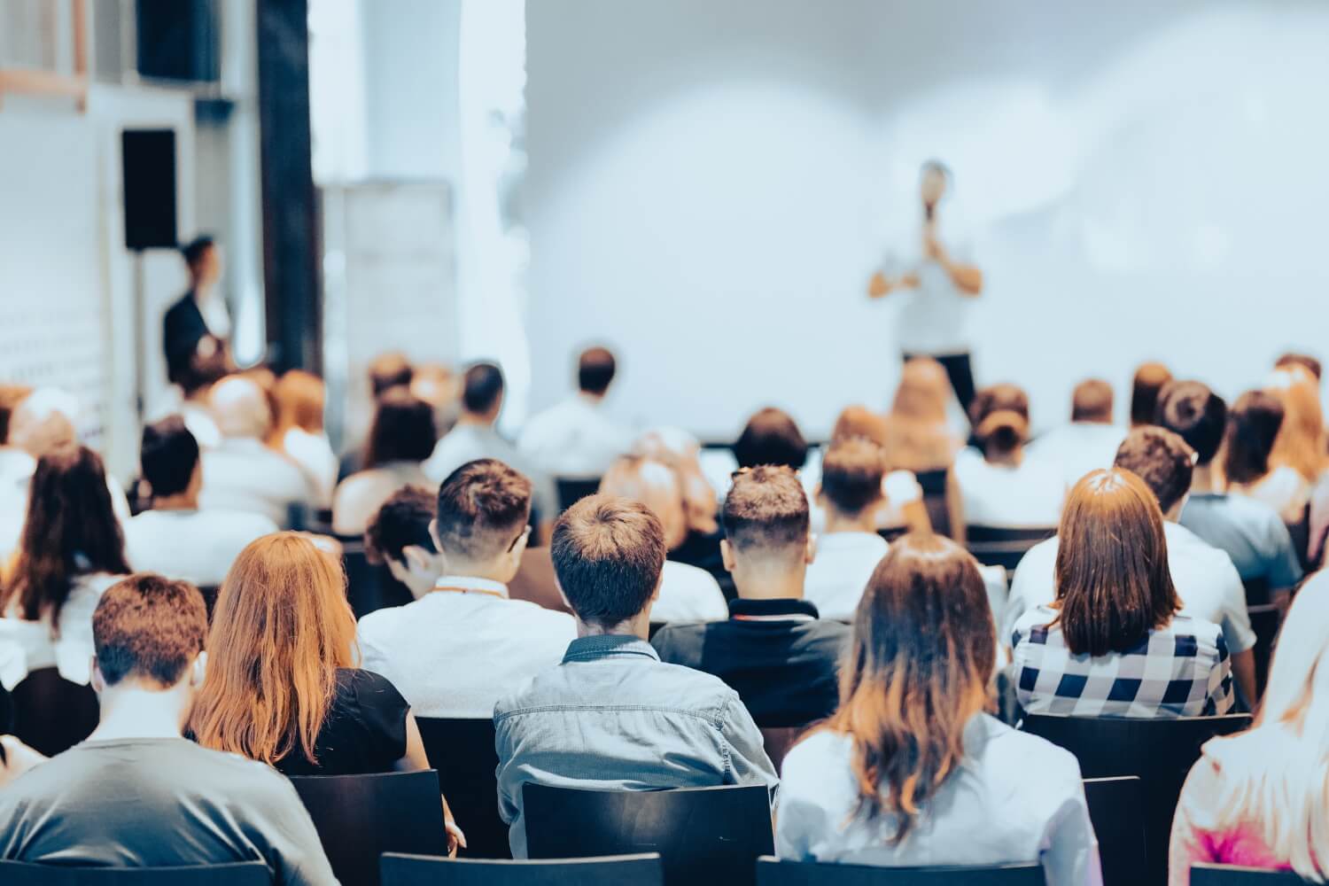 A large audience sits in rows of chairs, facing a person standing and presenting on conformal coatings technologies in front of a bright, white screen.